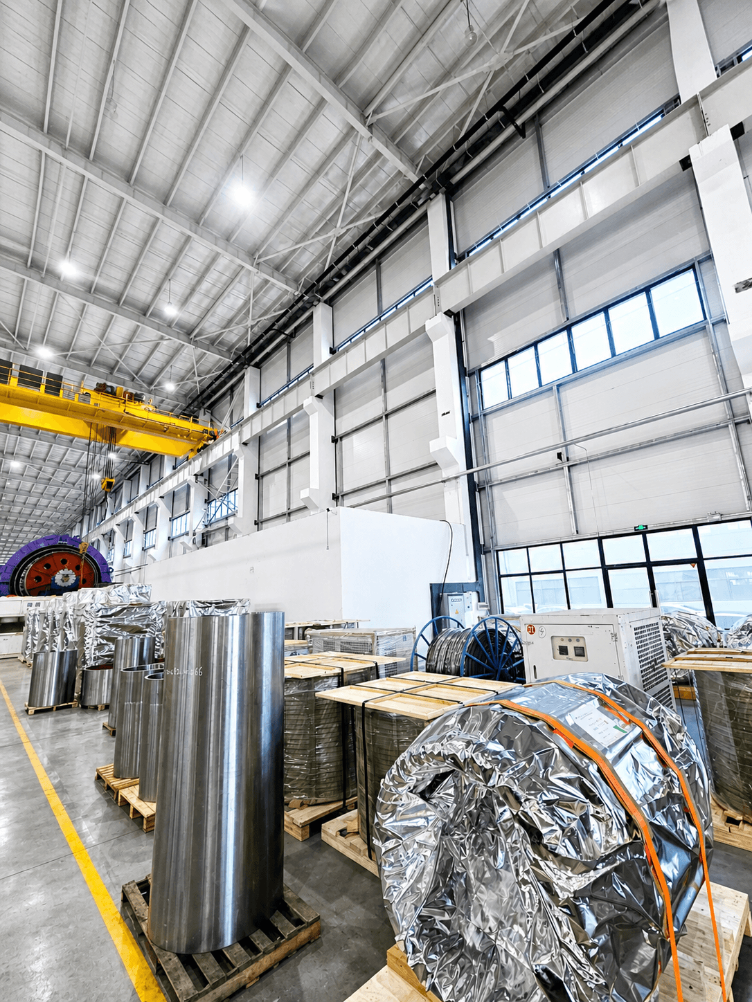 Large industrial warehouse interior with metal cylinders, foil-wrapped rolls, and a yellow overhead crane.